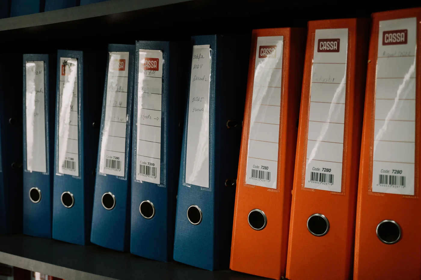 Archivadores de palanca azules y naranjos alineados en un estante; fotografía de Zulfugar Karimov en Pexels.